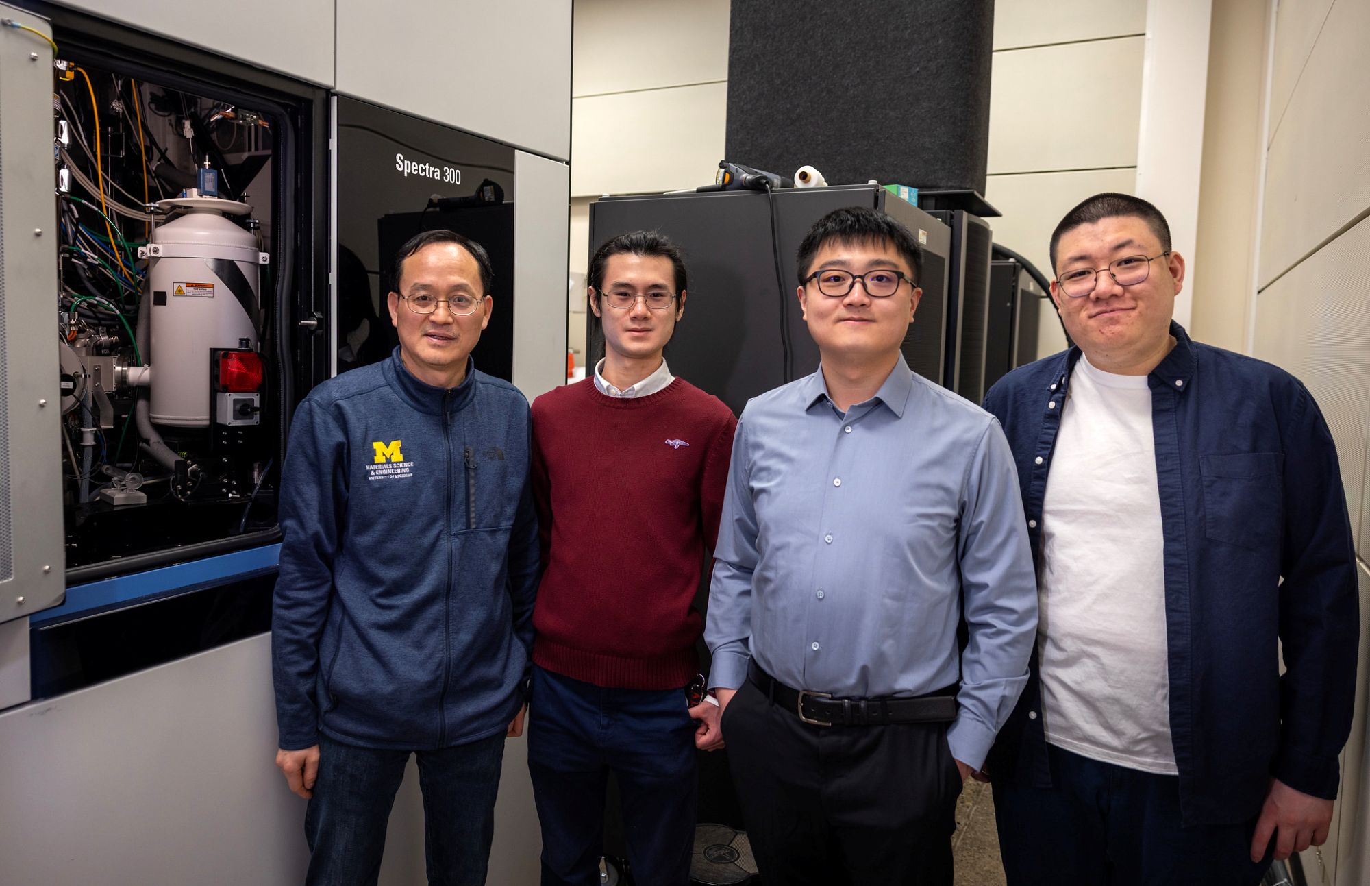 Four men stand near a window showcasing a cylindrical structure and many wires and tubes.