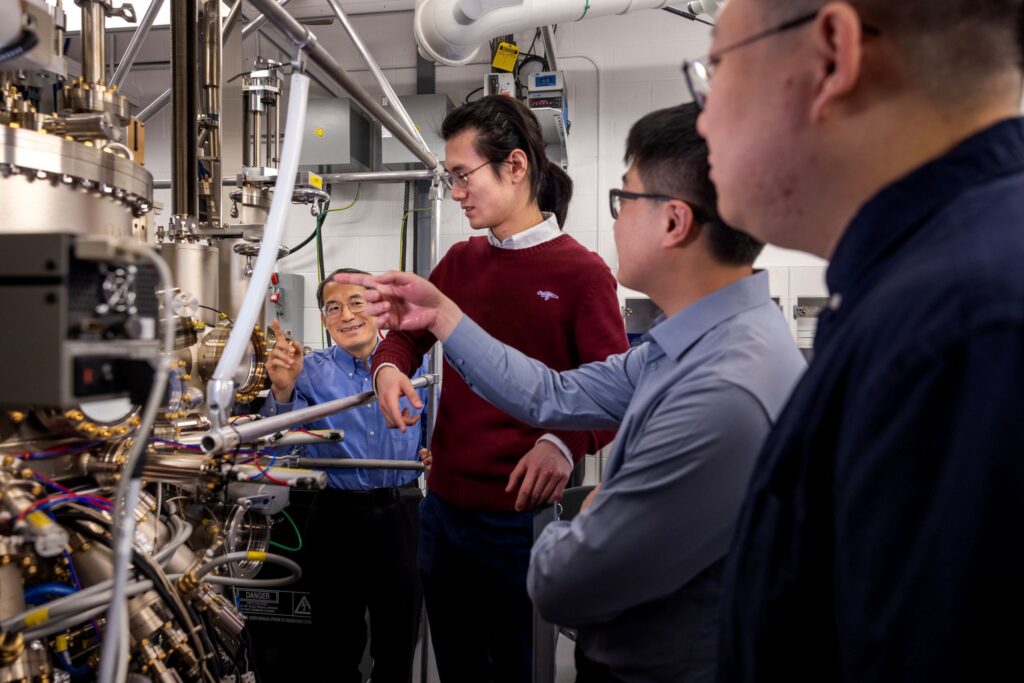 Four men stand in a line, diagonal toward the camera, pointing at a human-height silver and gold machine with many valves and tubes branching from it.