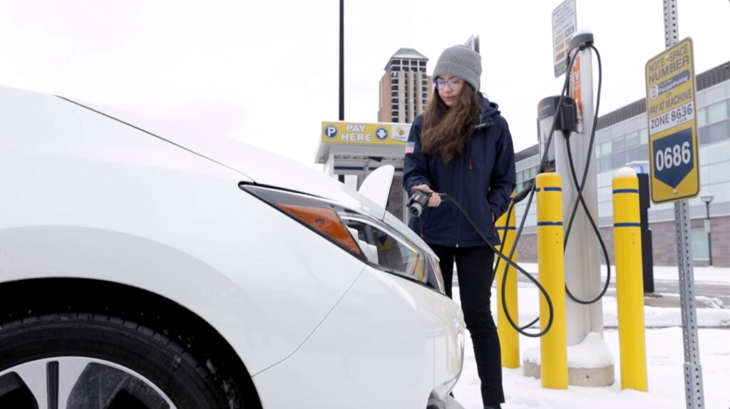 A woman, wearing a jacket and winter hat, prepares to connect an electric vehicle to a charger.