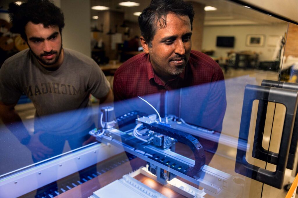 Two men are reflected in the glass of the 3D printer, which is producing a plastic structure.