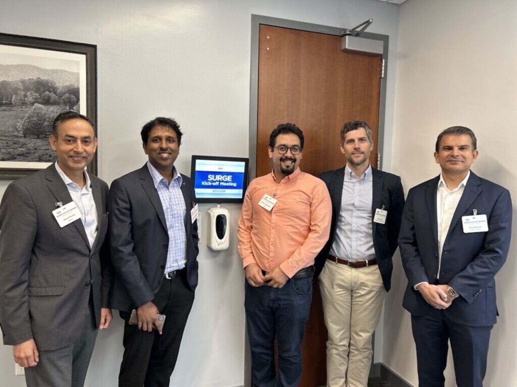 Five men stand near a meeting room door with a screen that reads "SURGE."