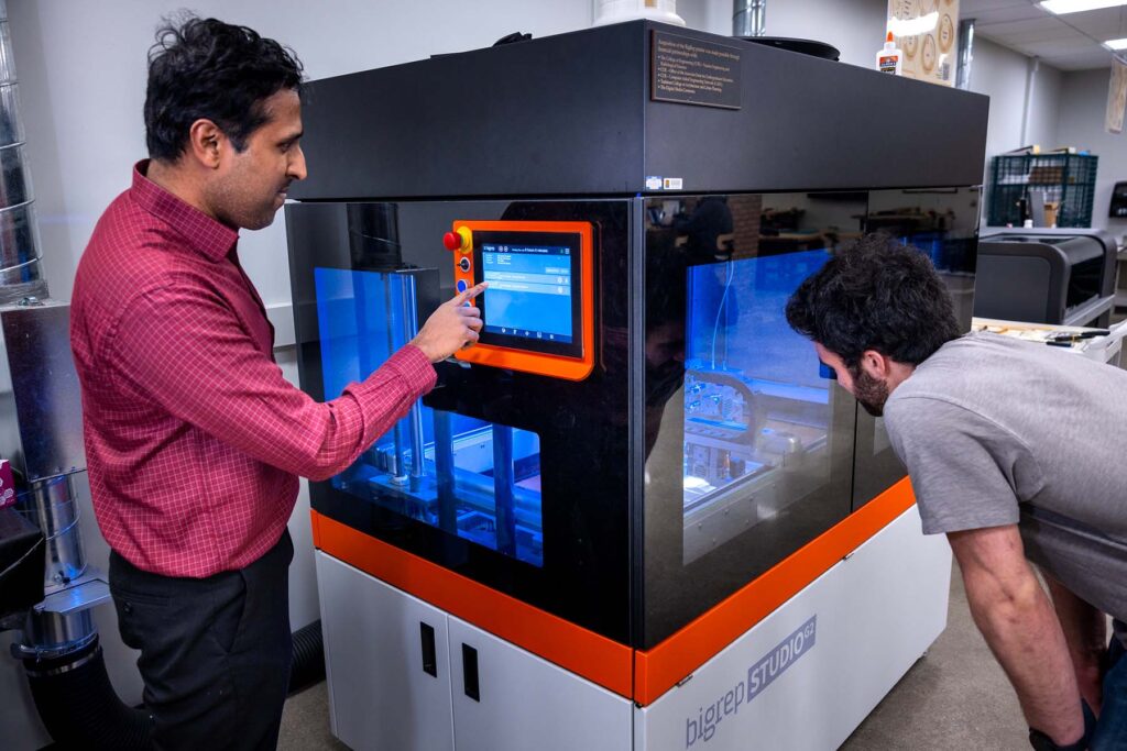 One man makes selections on the touch screen of the 3D printer while another peers into the glass case.