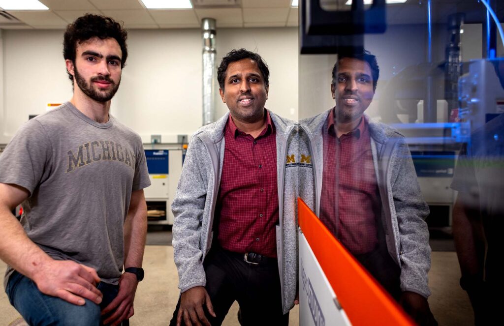 Two men are reflected in the glass of the 3D printer, which is producing a plastic structure.