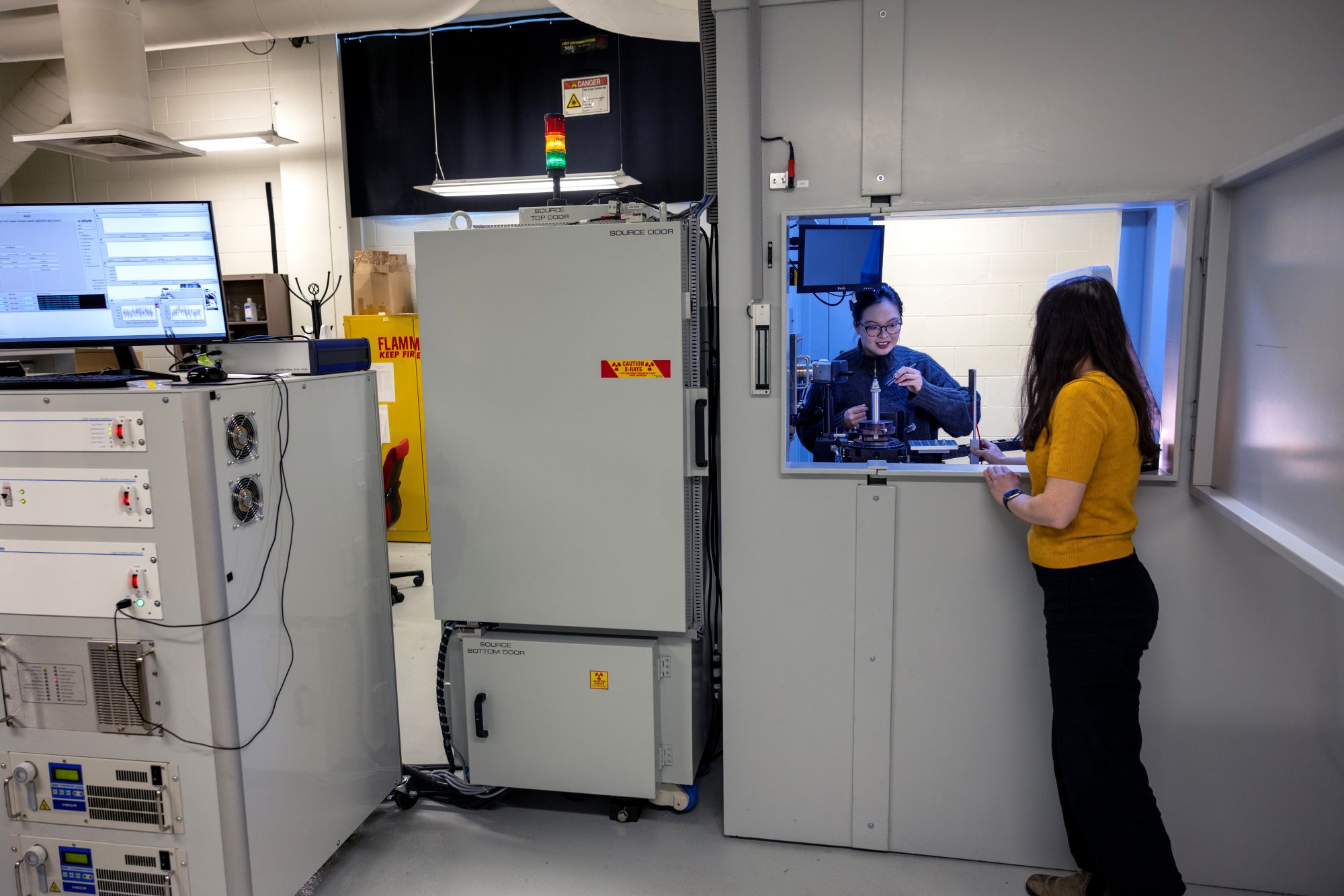 Two people work in a laboratory: In the background, an open window looks into a small room where one woman positions a sample on a microscope stand. In the foreground, a second woman stands in the larger room, observing through the window. Two refrigerator-sized devices stand to the left of the room with the microscope stand.