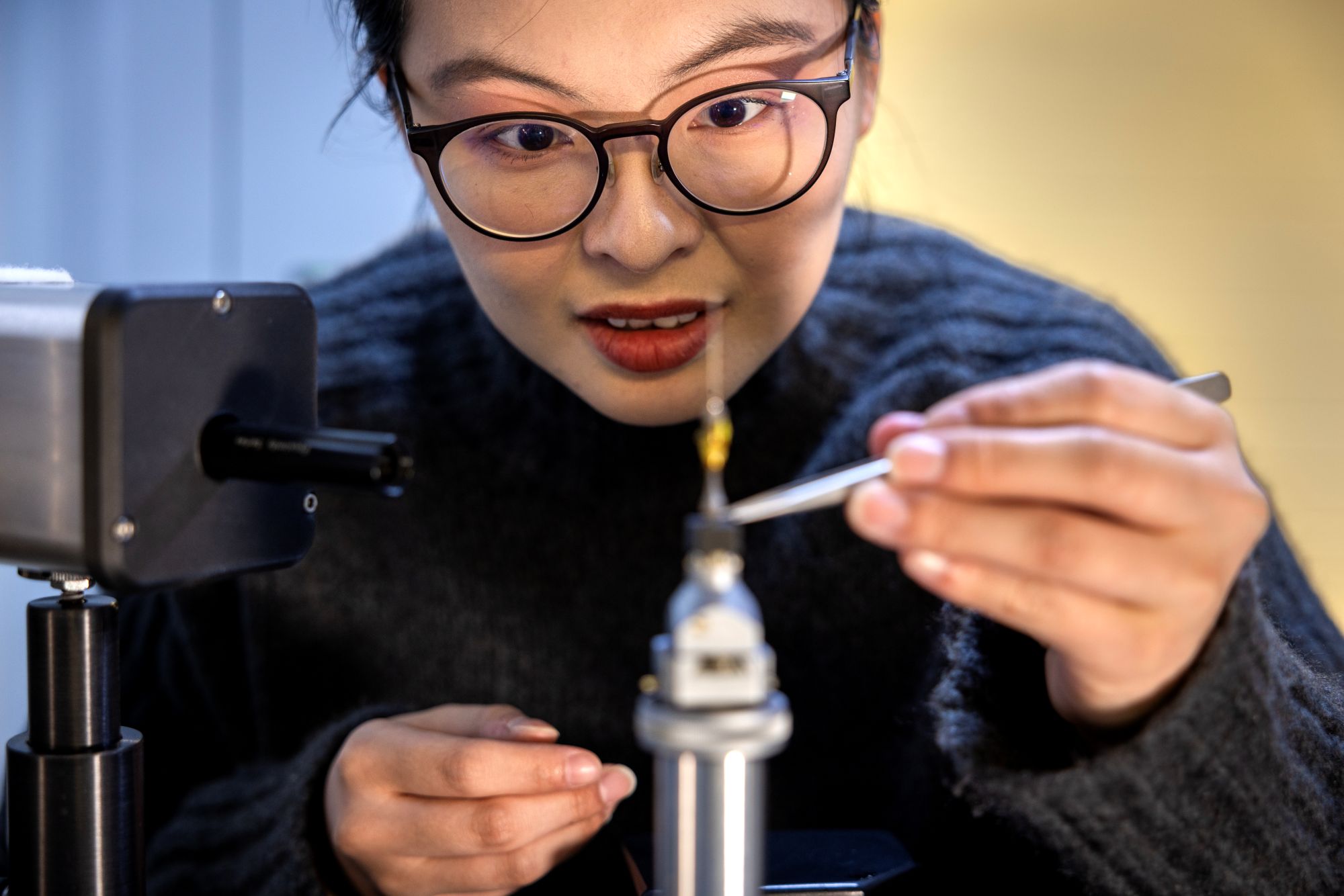 An in-focus woman in the background holds tweezers in the out-of-focus foreground as she positions a small metal rod on a silver, cylindrical microscope stand.