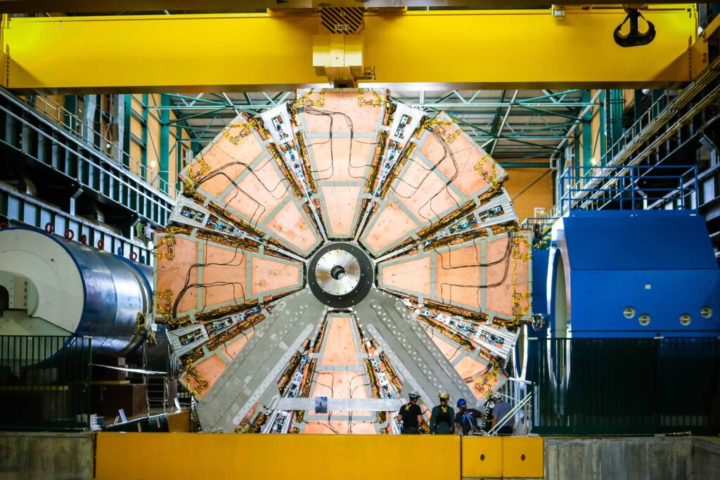 A large, circular section of the ATLAS detector at CERN’s Large Hadron Collider is shown, viewed head-on. The detector has a complex, mechanical appearance, with a segmented, pie-slice design featuring orange and metallic panels, wires, and machinery radiating from a central hub.