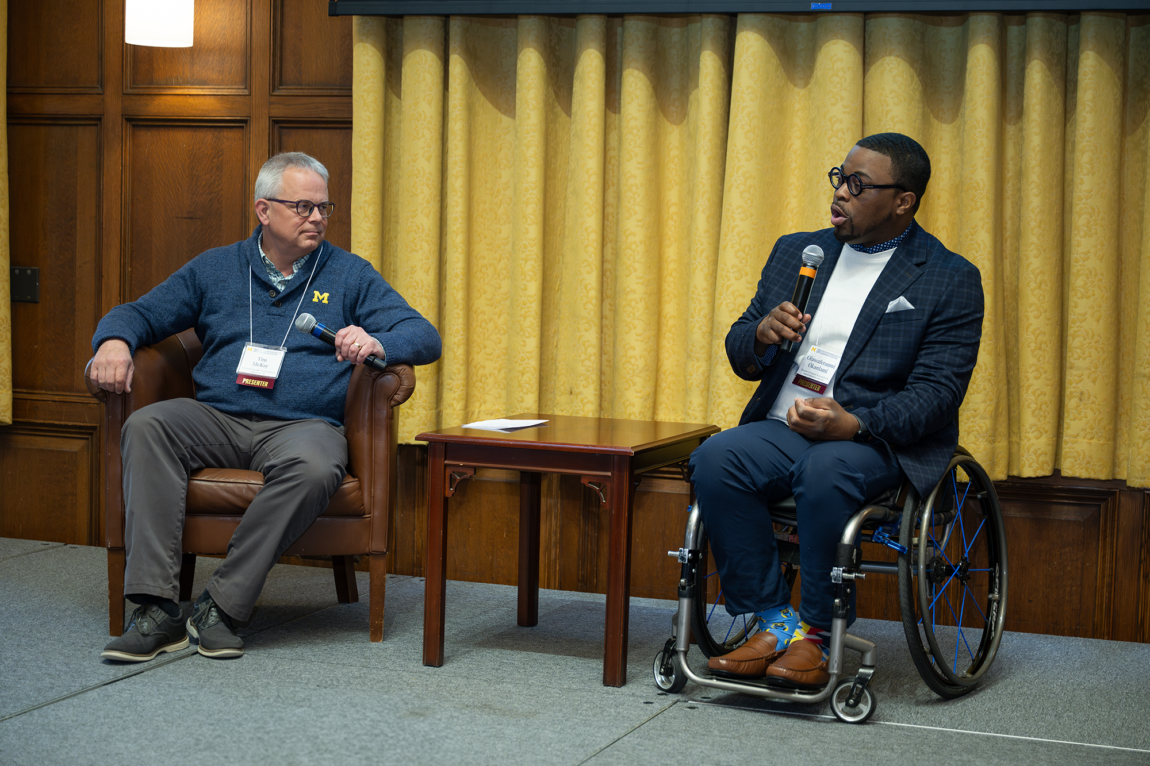 One person seated in a chair and another in a wheelchair talking into microphones