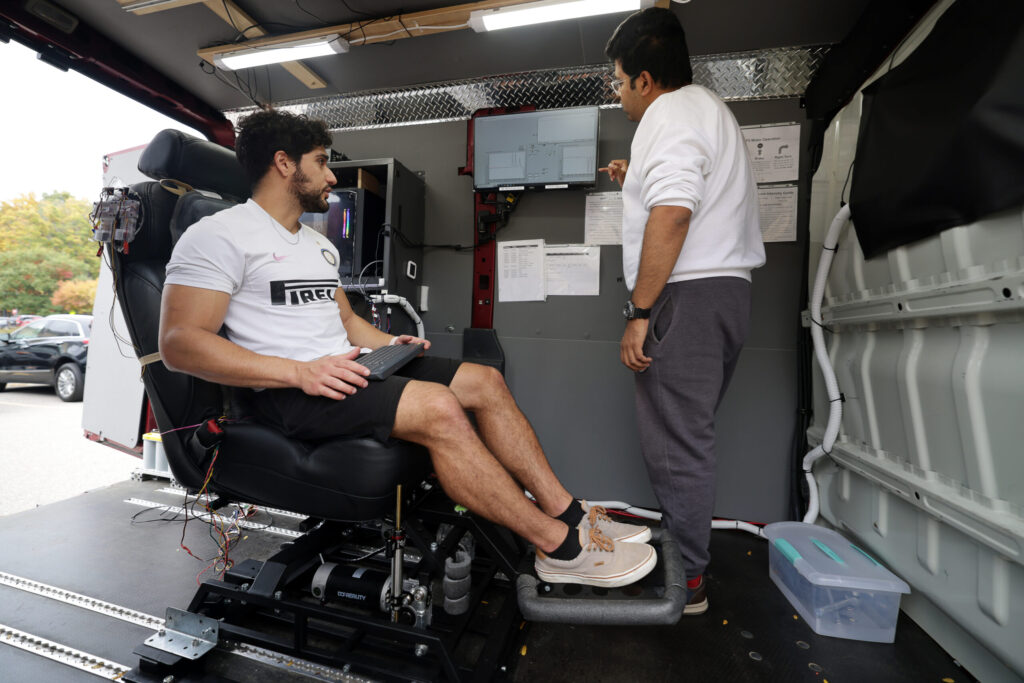 A man in a white t-shirt and shorts sits, at left, in a mechanized chair in the rear compartment of a van, while another man, standing, at right, analyzes data on a screen mounted to the van wall.