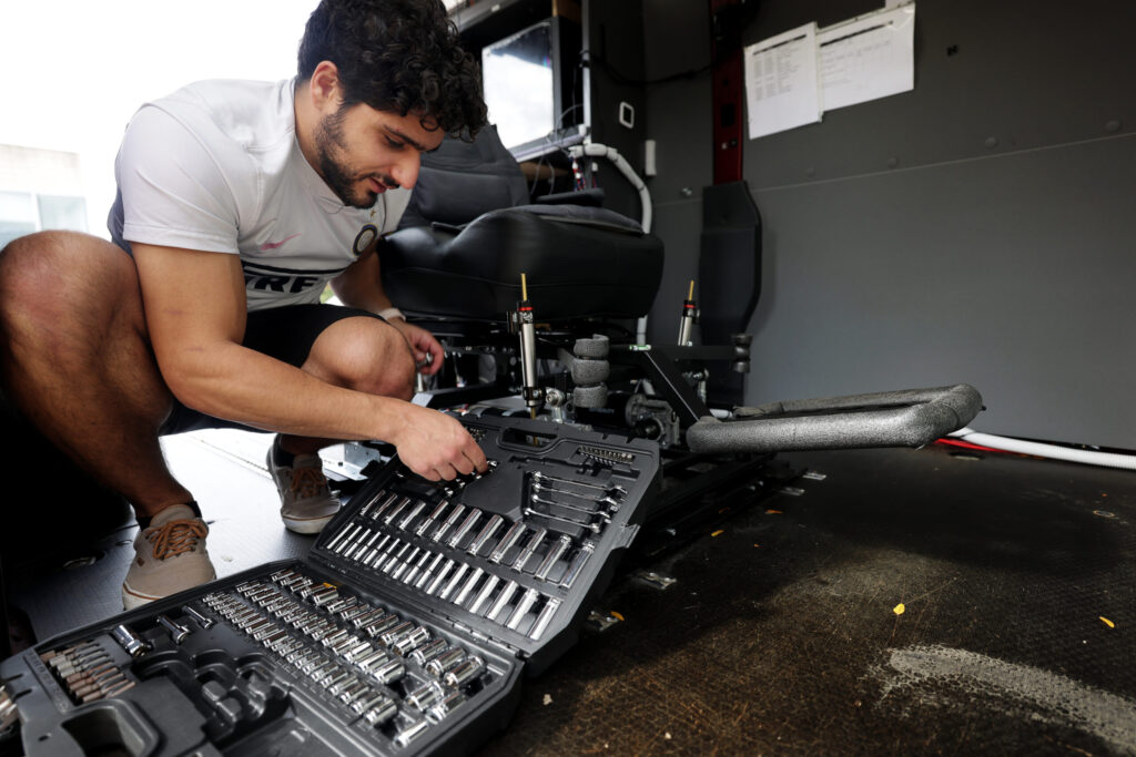A man, in shorts and a t-shirt, crouches next to a mechanized chair, with a tool kit laid out in the foreground. 