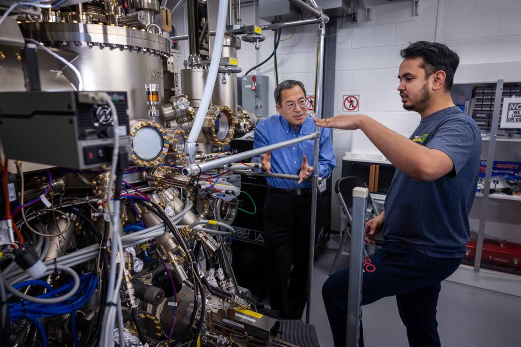 Two men stand facing each other in conversation, next to a large silver and gold machine with many tubes and valves branching off of it.