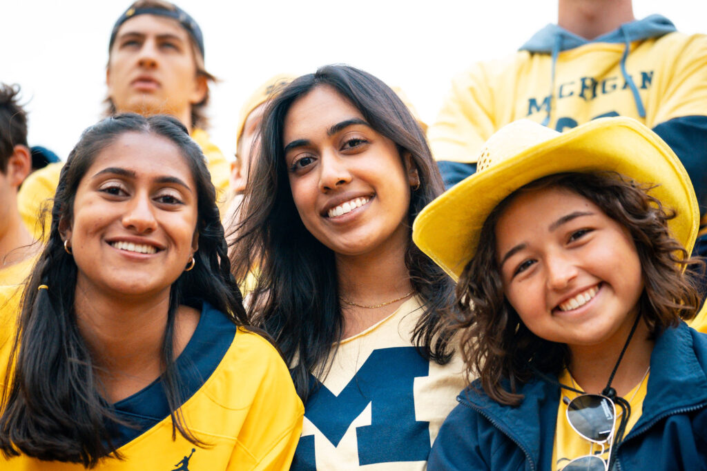 Three students pose for a photo with various pieces of Michigan clothing on, the person in the middle has a big Block M on their T-shirt. Image courtesy: U-M Center for Academic Innovation