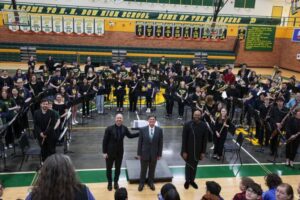 Jason Fettig, Stephen DeRees, music education teacher at H. H. Dow High School, and DaJuan Brooks, U-M doctoral student, with the combined college and high school student band. They are in a high school gym with green and yellow signage.
