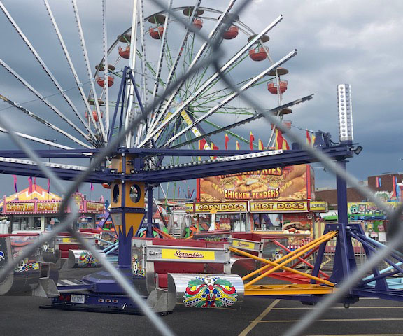 A view through the chain link fence of a carnival that hast yet to open. Stormy skies and a Ferris Wheel.