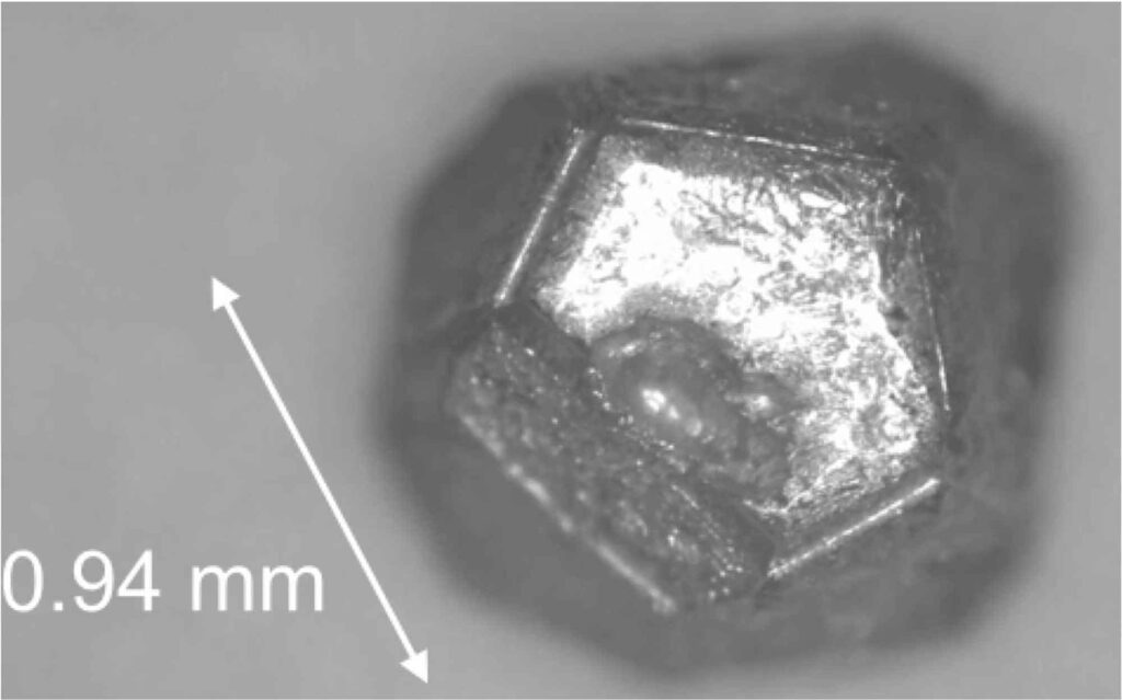 A silvery, metallic quasicrystal grain resembles a 12-sided die used in tabletop games. A scale bar shows the diameter of the grain is 0.94 millimeters.