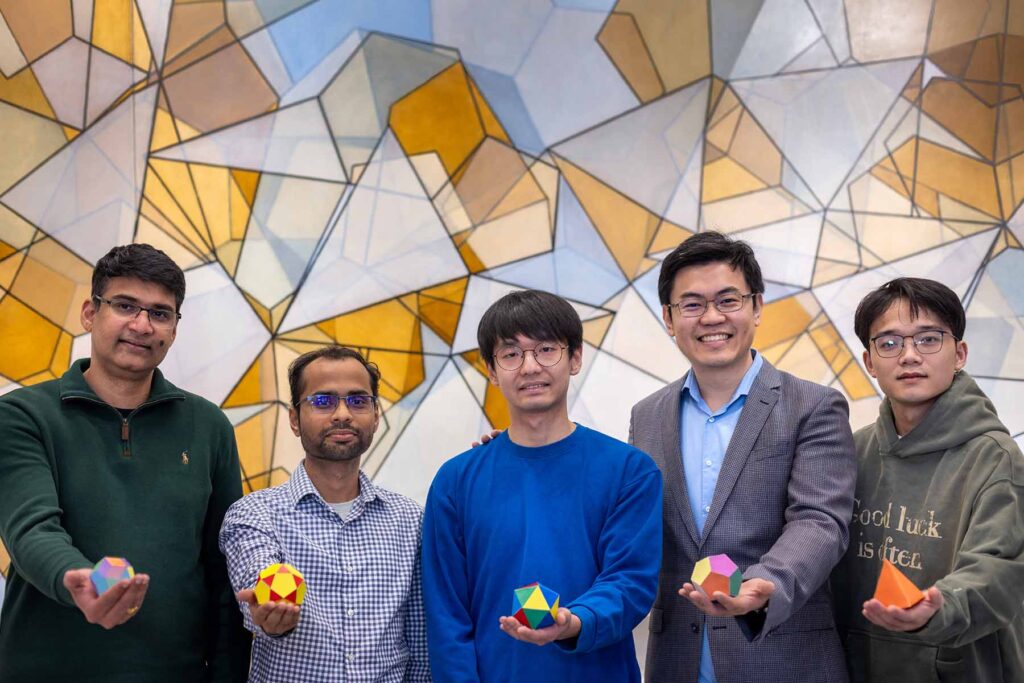 Five men stand in front of a wall, decorated with overlapping orange, gray and blue polygons. Each man holds a large die-like object. From left to right, the men hold: a crystal with rhombus-shaped faces; a crystal with pentagonal and triangular faces; a crystal with 20 triangular faces; a crystal with 12 pentagonal faces, and a four-faced pyramid.