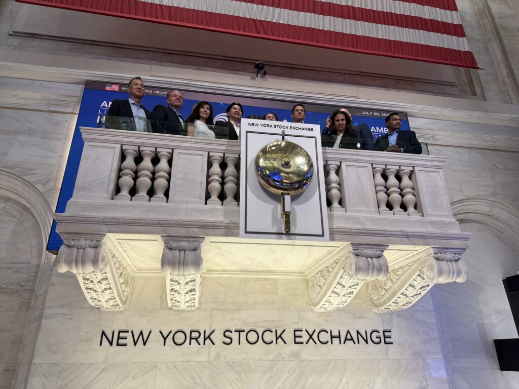 Men and women dressed in suits stand on a balcony behind a large gold bell. Below the balcony, lettering on the wall says New York Stock Exchange.