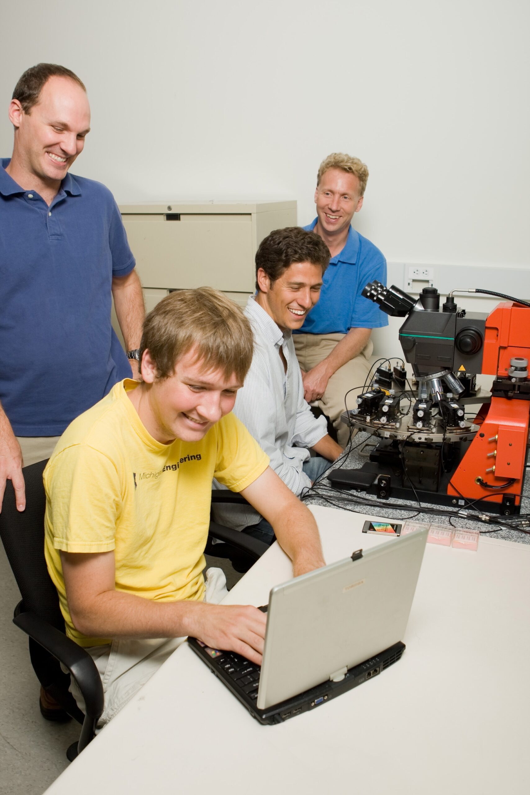 A man in a yellow shirt works at a computer while another sits at a device that looks like a complicated microscope. Two other men in blue shirts look over their shoulders.