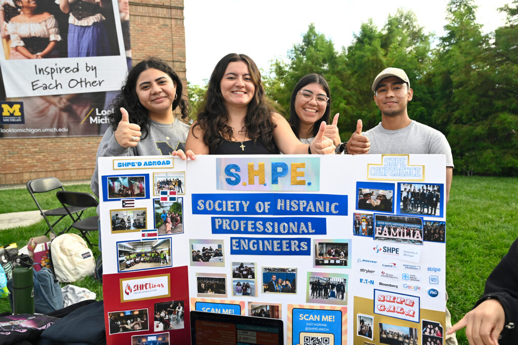 Four people stand behind a posterbooard and hold up their thumbs