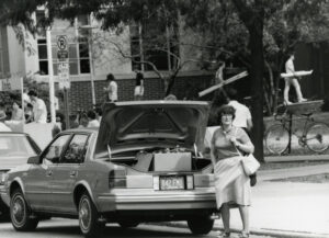 Black and white image, circa 1980, of woman standing by car with open trunk as the frenzy of move-in day unfolds around her.