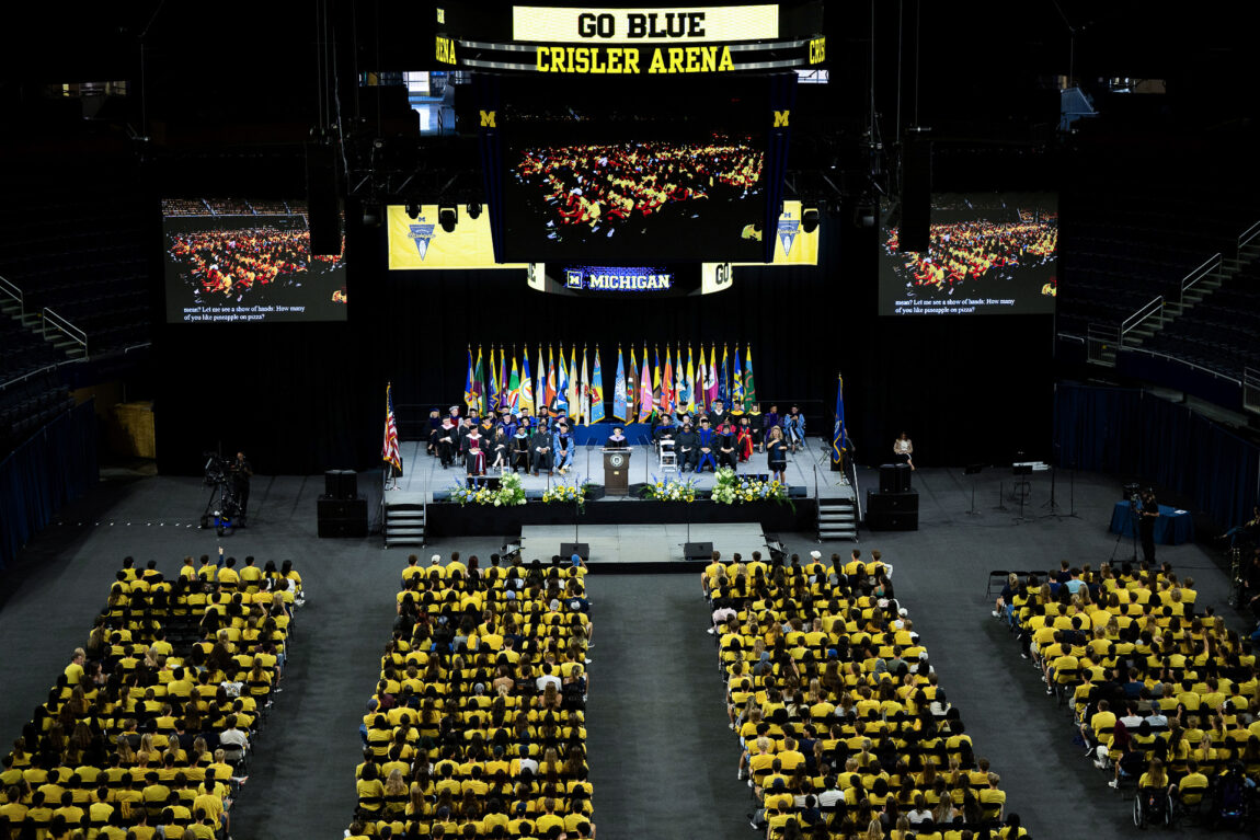 An overhead view of a gathering of people, with seating in four sections and others sitting on a stage