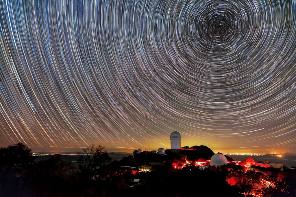 Whirling lines in the sky are the trails of stars after an hour-long exposure above Kitt Peak National Observatory. Different colors in the trails show the different temperatures of the stars, with blue being the hottest stars and yellow/red the coolest. A The Nicholas U. Mayall 4-meter Telescope is visible above the horizon, and a red glow appears on the mountain, caused by red lights used to ensure the eyes of visitors and staff remain dark adapted at night. There's a yellow glow above the horizon, which comes from Tucson, AZ, about 89 kilometers (55 miles) to the northeast.