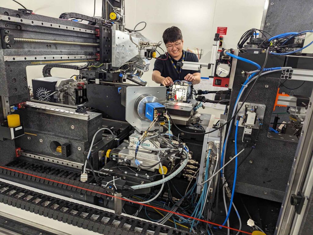 n the foreground, a large metal piece of equipment with many wires and sensors protruding. In the background, a man smiles while placing an object on the instrument.