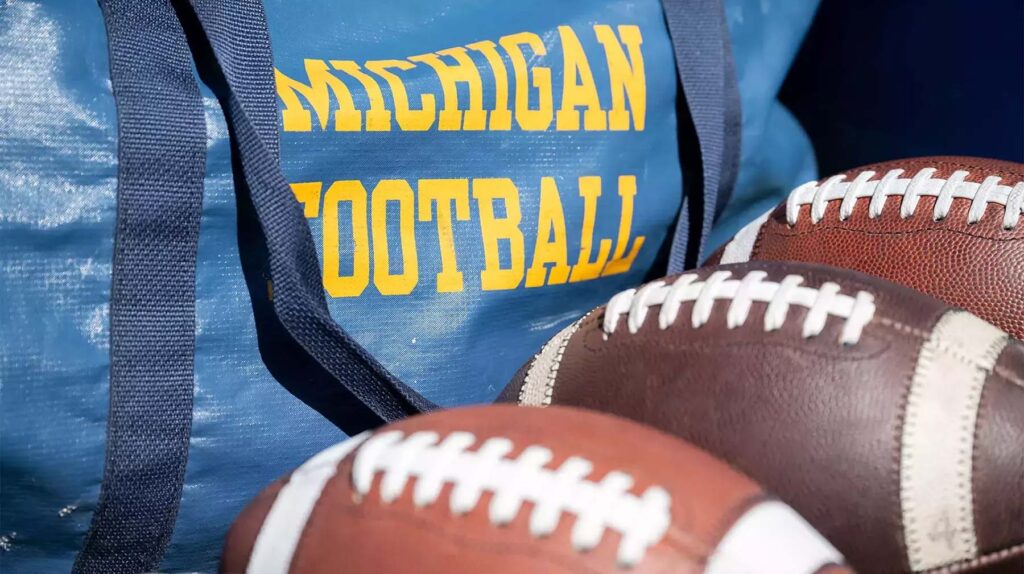 Close-up of a duffel bag that says "Michigan Football" with three footballs in the foreground. Image credit: Michigan Photography
