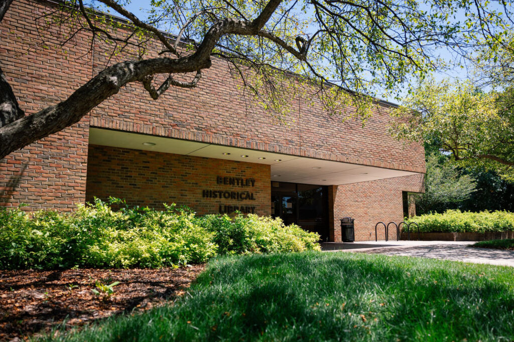 A tree and garden of small bushes sit in front of the one story, light brown brick building hosing the Bentley Historical Library. The doorway and mounted letters reading “Bentley Historical Library” sit shaded under a large overhang.