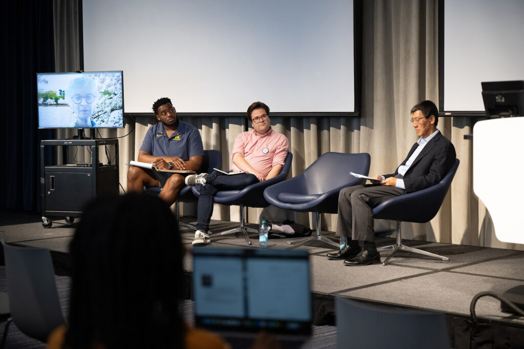Three people sit in chairs while another listens on a computer screen during an event