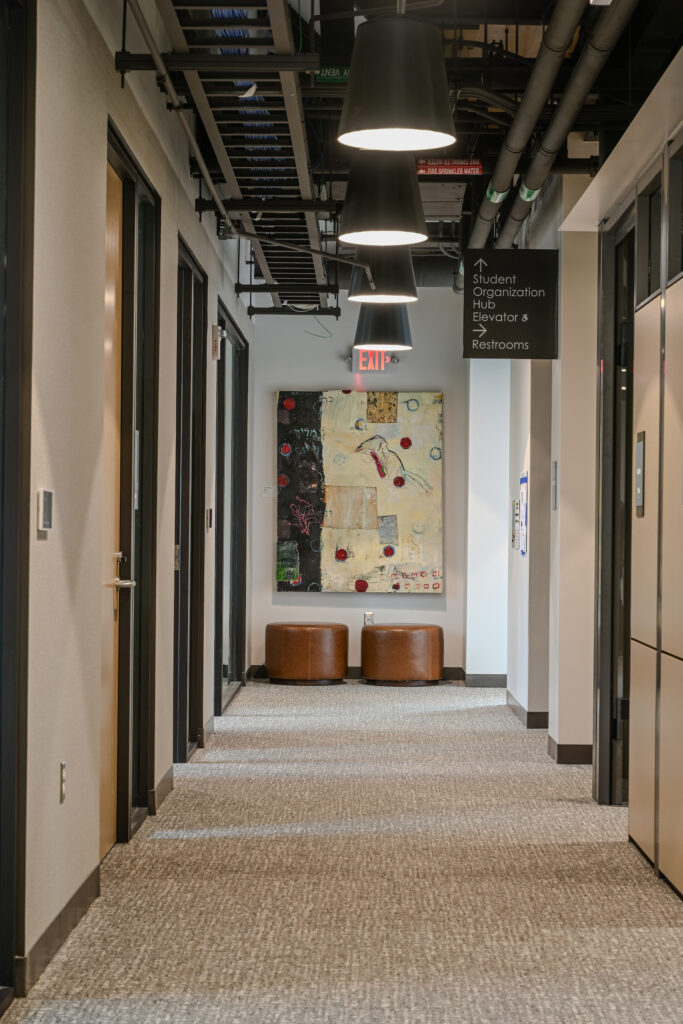 A long hallway at the end of which sits two brown cushions on the floor and a colorful art display on the wall