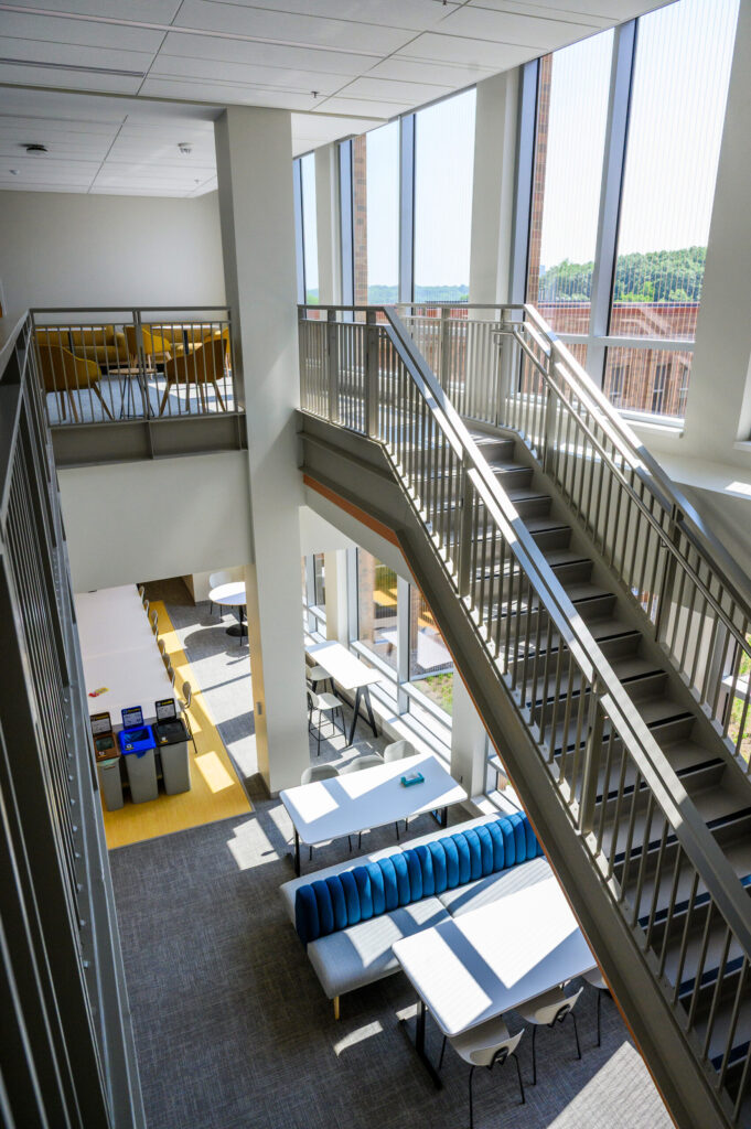 A look from above of a kitchen and lounge area of a building with multiple tables and seating options and a staircase to the right