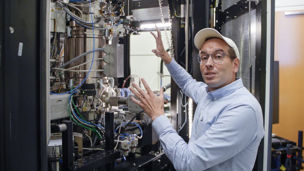 A man gestures toward a complex network of wires, plugs and metal pipes inside a cabinet-like container.