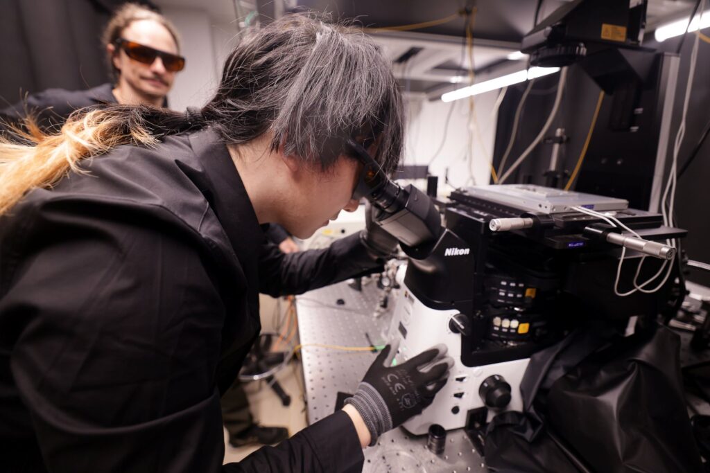 Jiang leans over the black eyepieces, dark-lensed laser safety glasses on. Florian also wears laser safety glasses.
