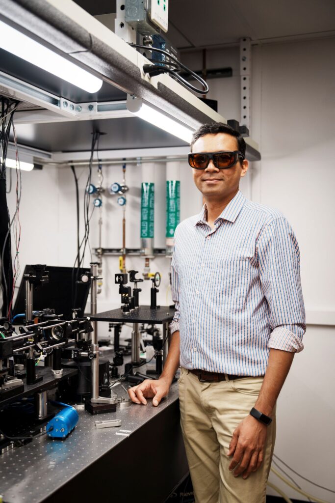 Parag Deotare stands under a set of fluorescent lights, next to an optical table equipped with lasers, detectors and many lenses and mirrors, at different heights. He wears a pair of tinted laser safety glasses.