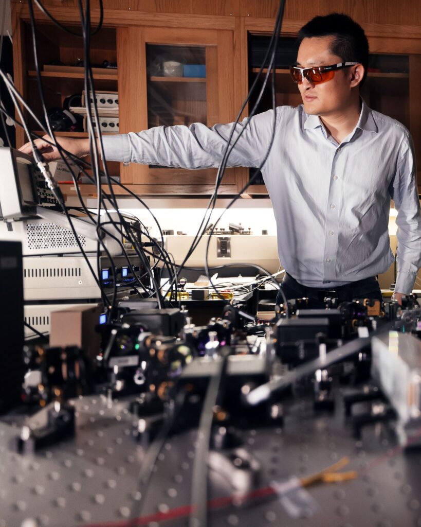 Zheshen Zhang adjusts a dial on a small screen with his arm outstretched, standing behind an optical table with a series of lenses. He wears a pair of tinted safety glasses.