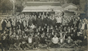 Black and white image, dated 1911, of a group of University of Michigan sophomores surrounding face-painted freshman captured while putting up posters in the 1908 "rush" hostilities.