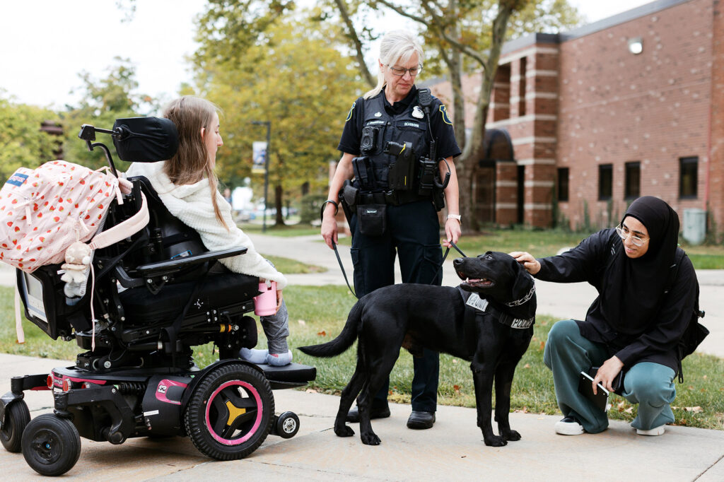A black police dog accompanied by an officer receives attention from two people on a sidewalk