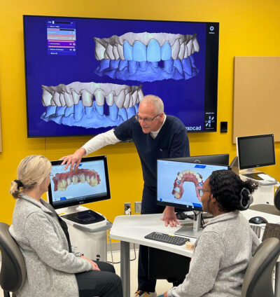 A man standing points at an image of teeth on a monitor while two women seated look on