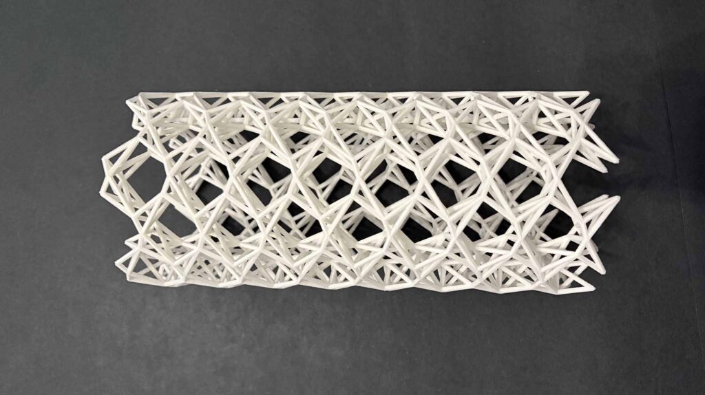 A white 3-D printed object stands on a dark background. Overall, it's fair to call the structure a tube, but it's made of an intricate, woven structure of twisting triangular and hexagonal subunits.