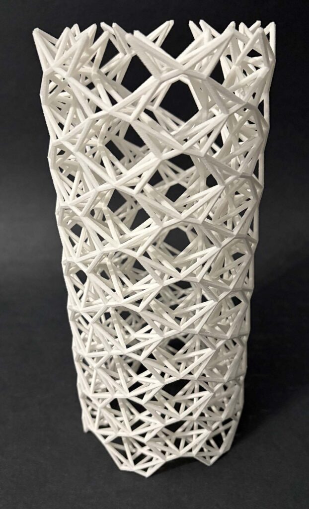 A white 3-D printed object stands on a dark background. Overall, it's fair to call the structure a tube, but it's made of an intricate, woven structure of twisting triangular and hexagonal subunits.