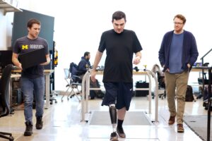 Three men walk through a lab with a treadmill built into the floor behind them. In the center, Seelhoff wears a prosthetic leg that stores its electronics inside a slim calf-shaped casing. On the left, Kevin carries an open laptop computer.