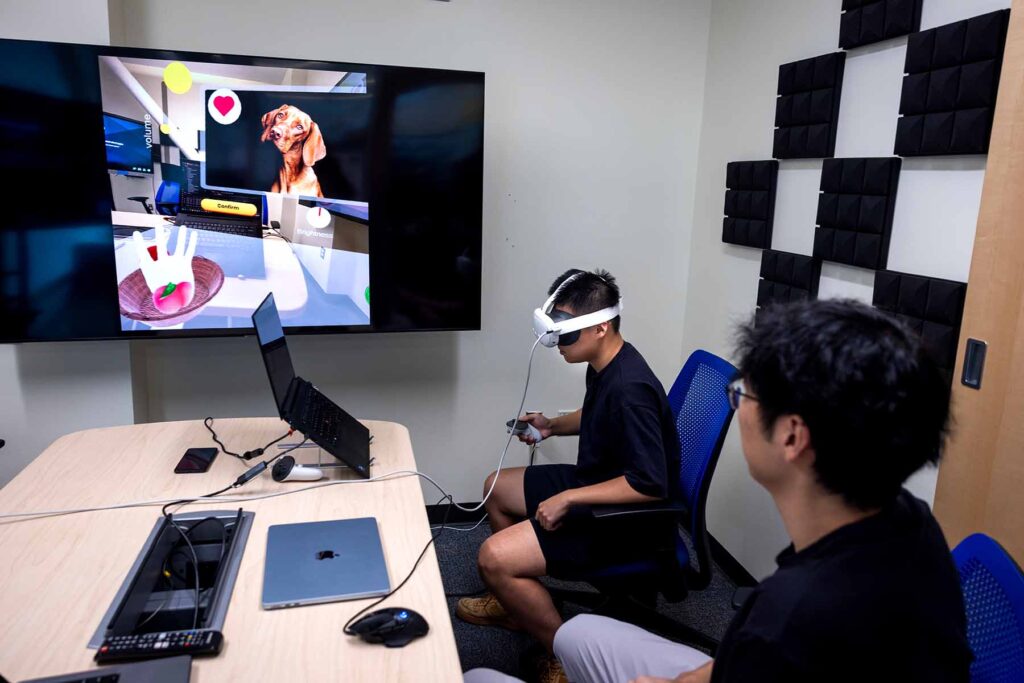 Two men sit at a video conference desk. One of the men is wearing a virtual-reality headset connected to an open laptop. A TV on the wall displays a white, disembodied hand hovering over a digital basket, which contains a digital apple. A photo of a dog is also displayed in the top right corner of the app window.