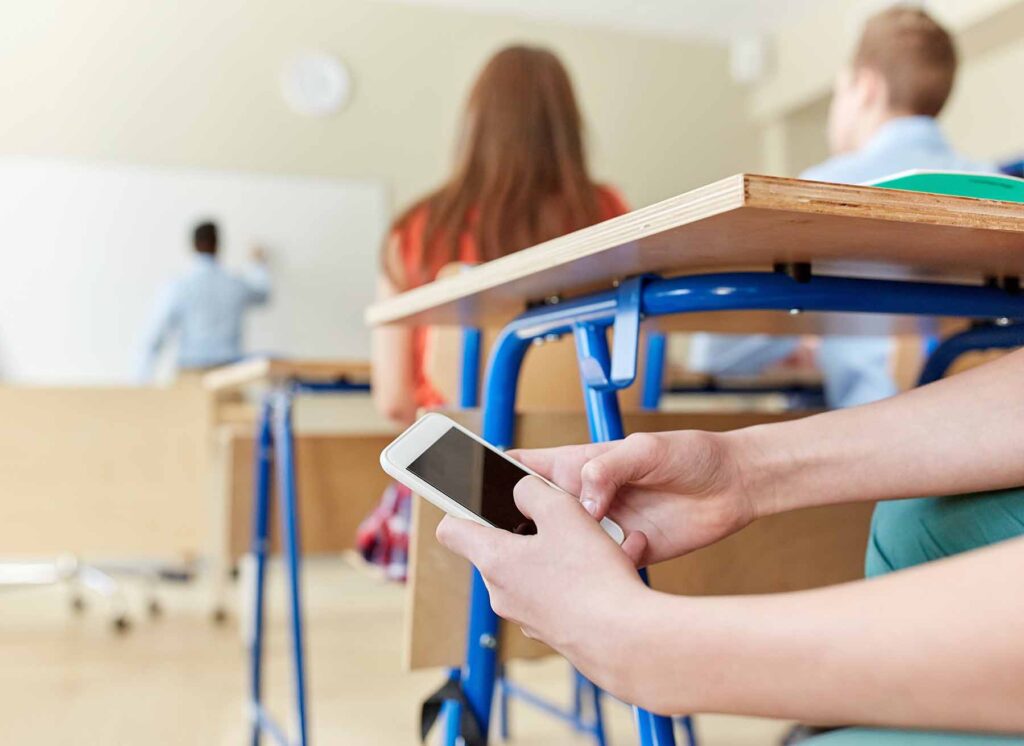 A student holds a cell phone under a desk in class. Image credit: Adobe Stock