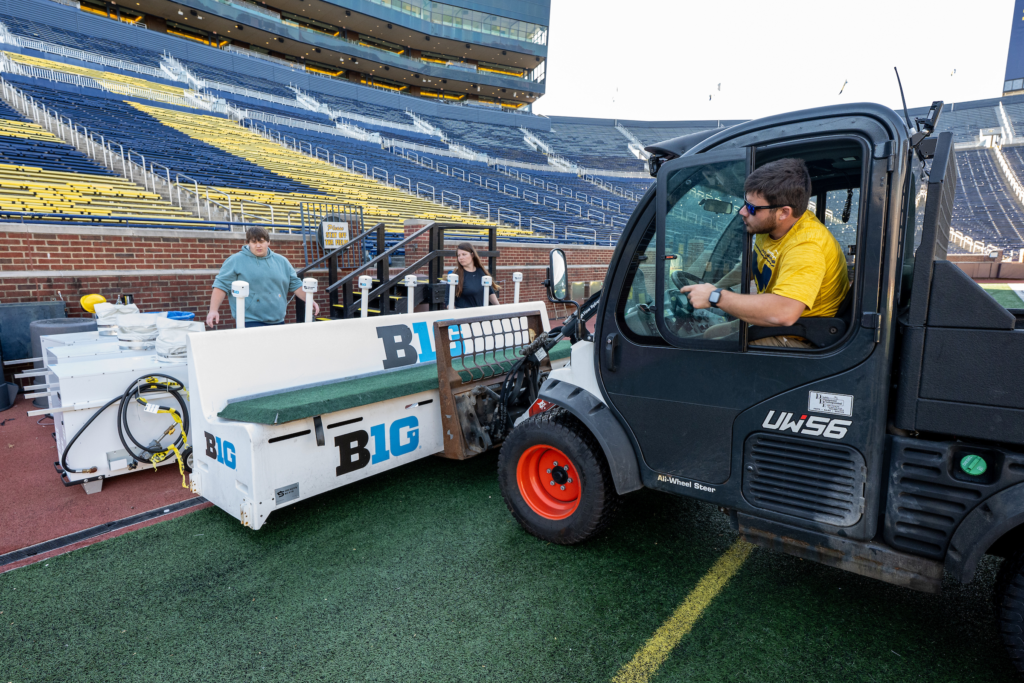 A person driving a vehicle places benches on the sideline of a field while two people stand nearby