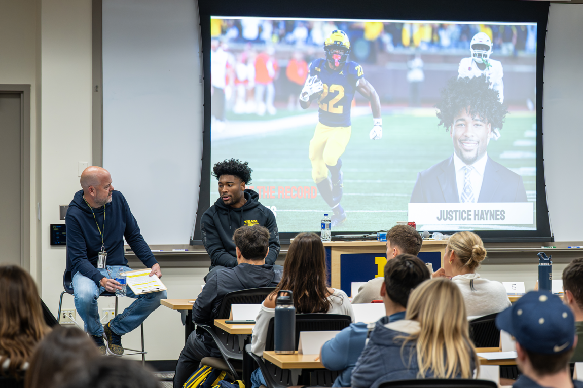Two men sitting in chairs speak to students sitting at desks