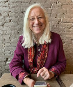 Smiling caucasian woman with white hair and glasses sits against a brick wall, dressed in vibrant colors.