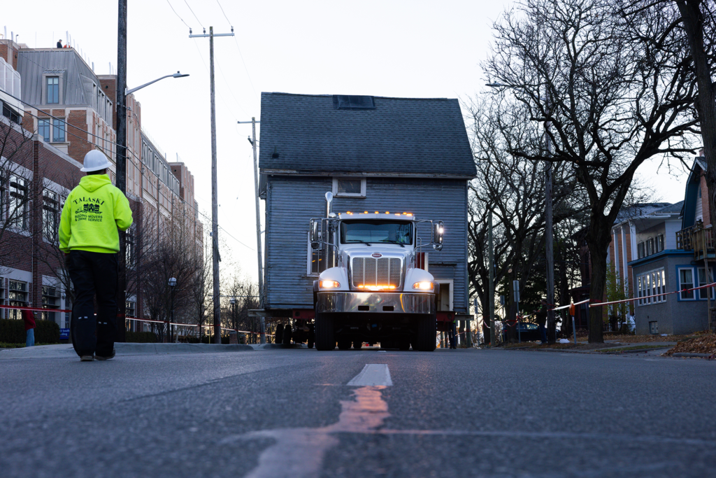a house is hauled down a street by a trailer while a worker in lime green stands nearby