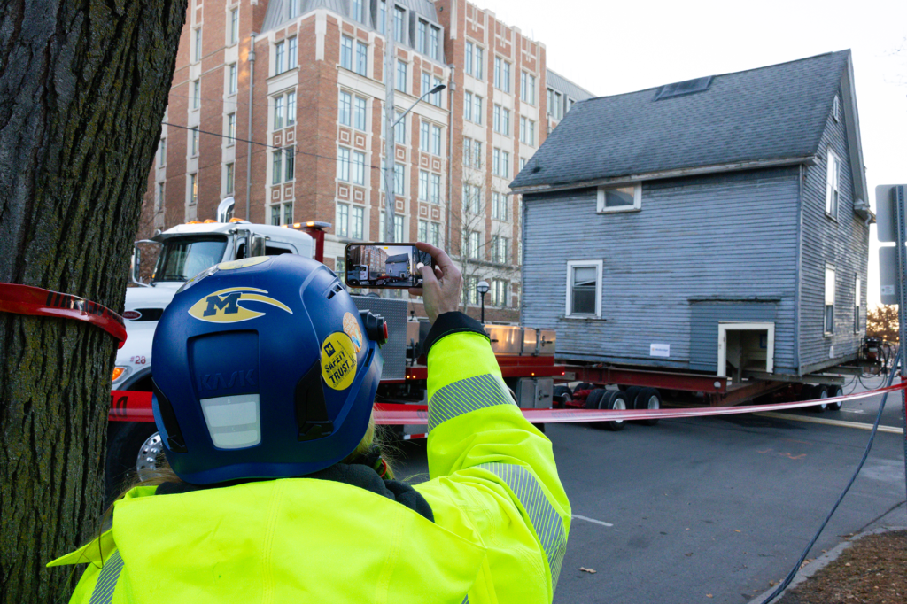 A person uses their phone to record a house being hauled down a street