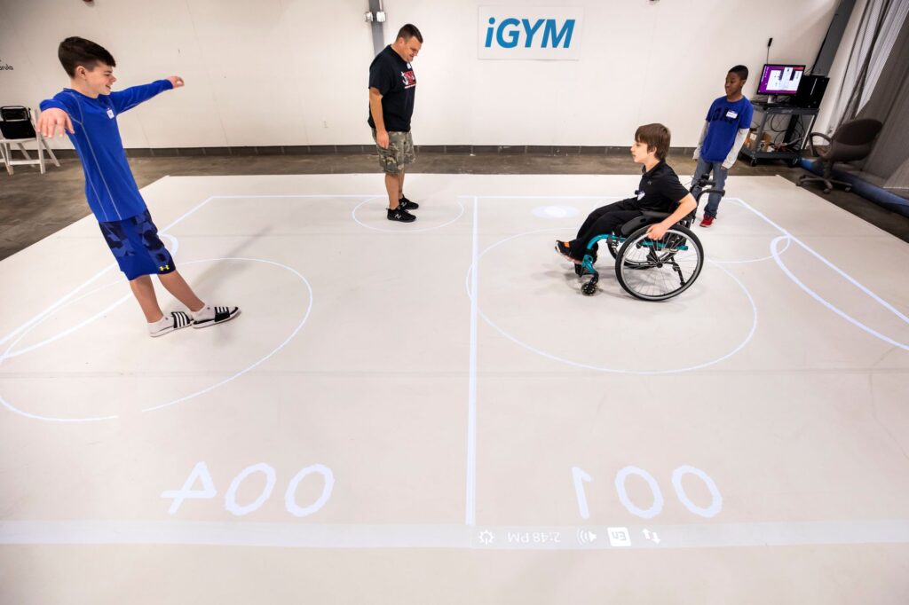 On the right, a boy grips the handrims of his wheelchair, the ball appearing as a white spot to his right. On the left, a boy stands with his arms outstretched and one foot in front of the other. The lines from the projector bound a rectangle around them and the center line between them as well as arcs marking the goals.