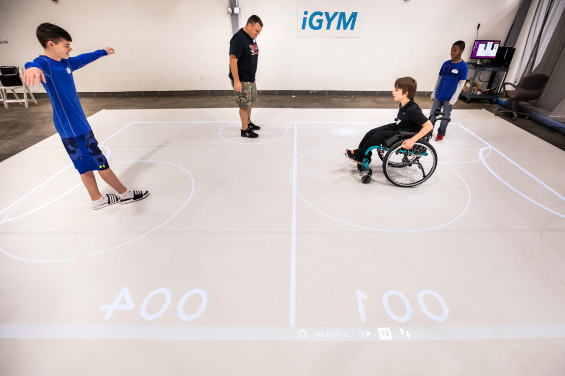 On the right, a boy grips the handrims of his wheelchair, the ball appearing as a white spot to his right. On the left, a boy stands with his arms outstretched and one foot in front of the other.